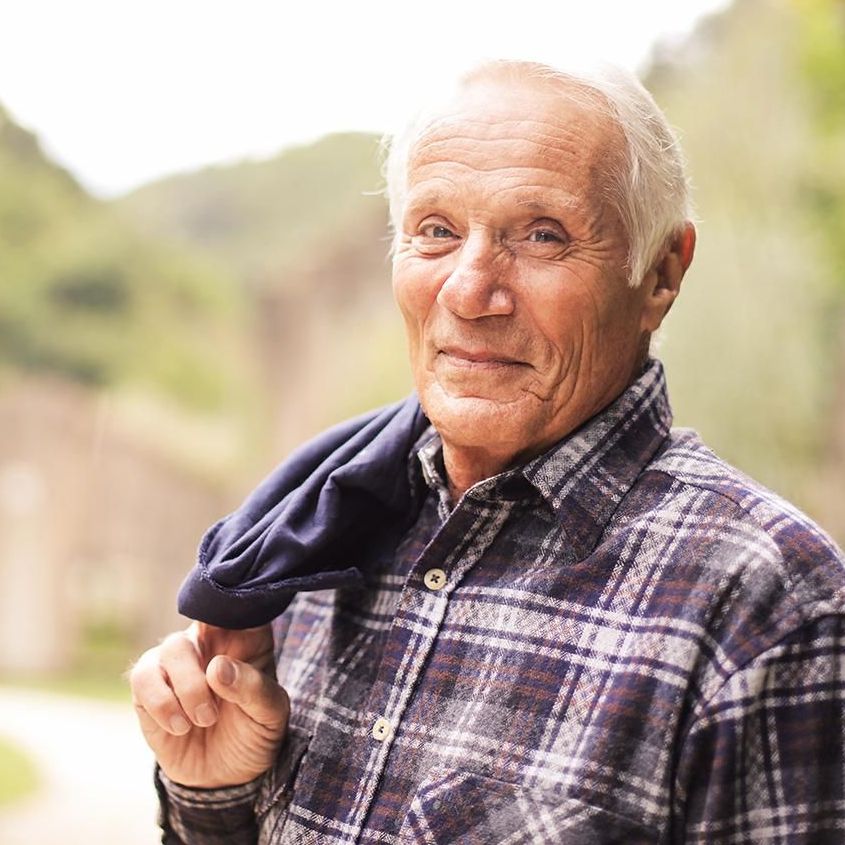 Older man with white hair wearing a plaid button up is smiling with his lips closed.